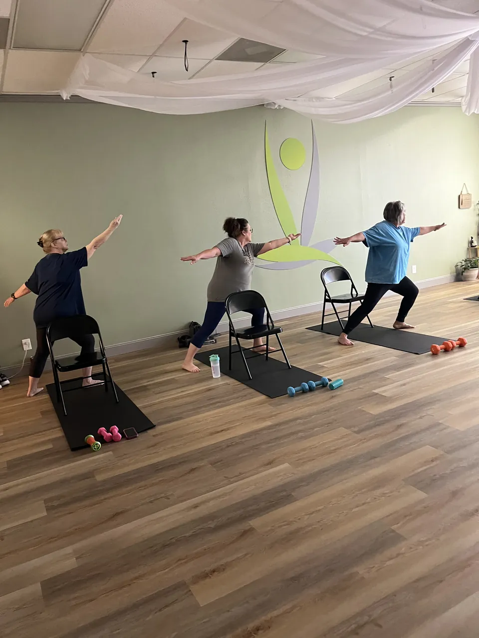 Three students in warrior two, each with a folding chair and hand weights on their mat, in front of the studio logo.