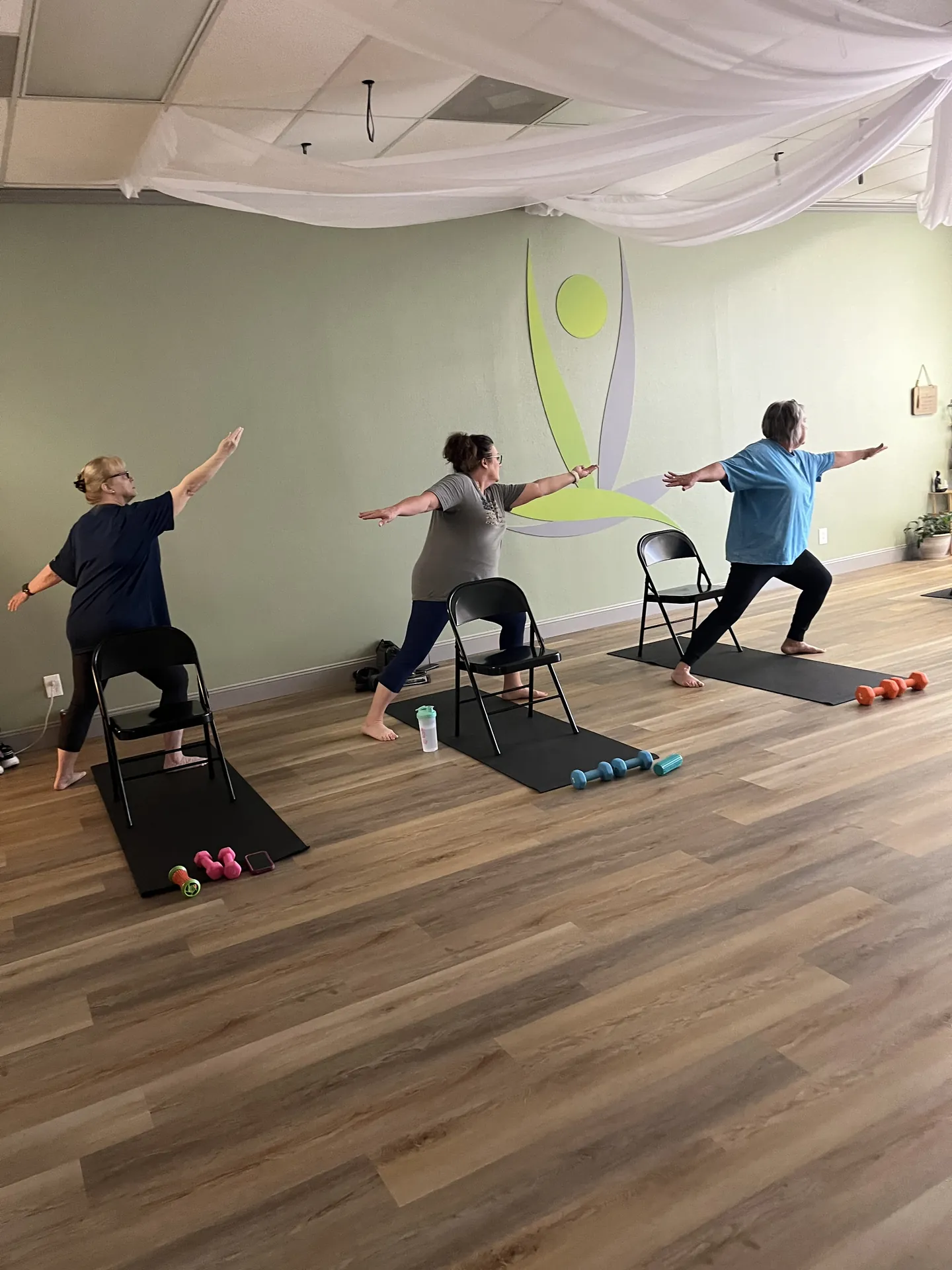Three students in warrior two, each with a folding chair and hand weights on their mat, in front of the studio logo.
